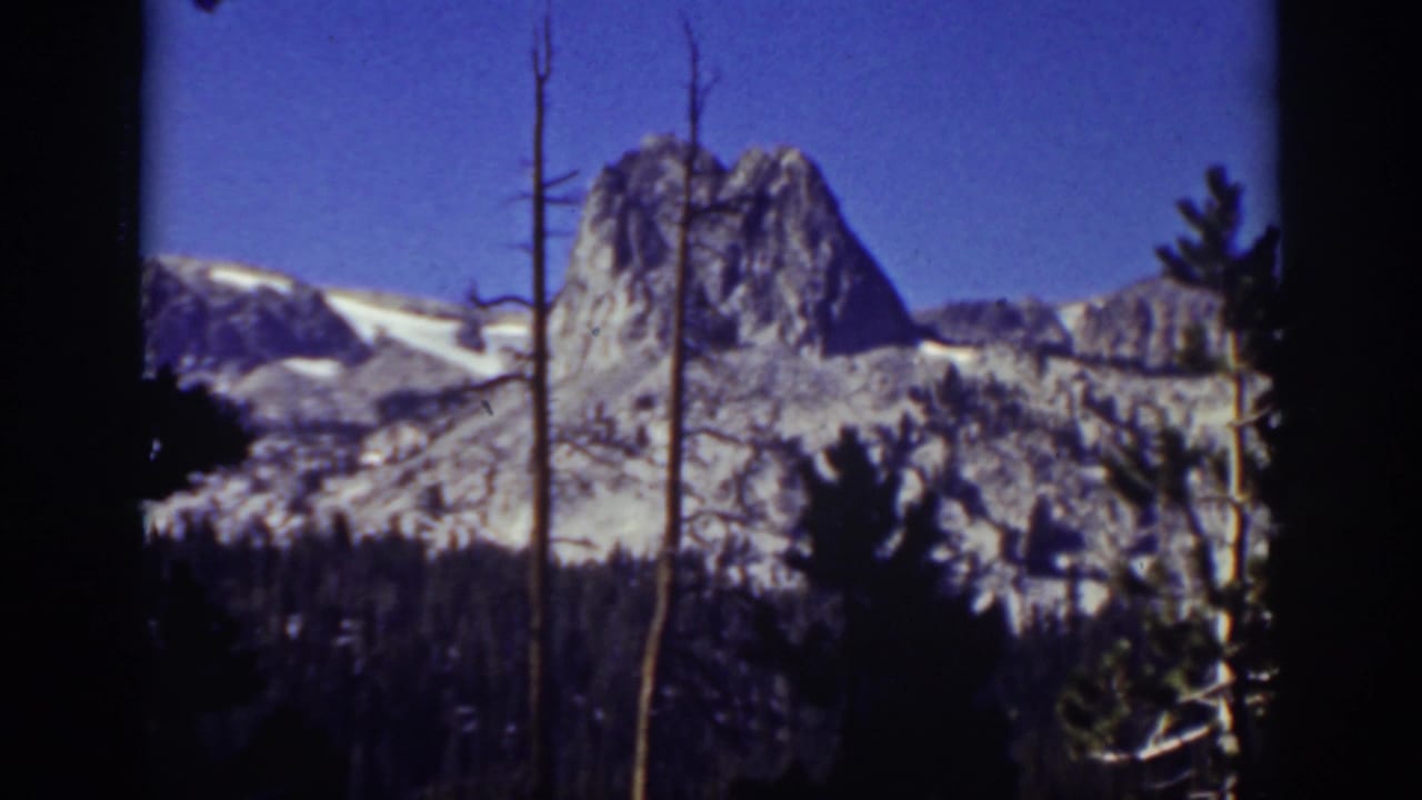 1939 Lake Tahoe 8mm frame showing a rocky Sierra Nevada peak above dark trees and bare trunks beneath a deep blue sky.