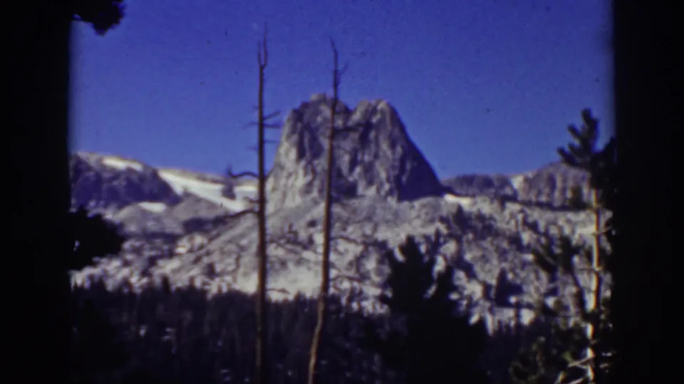 Lake Tahoe home-movie frame from 1939 with a snow-dusted mountain ridge, pine silhouettes, and bare trunks in the foreground.
