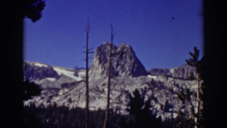 1939 Lake Tahoe mountain view on 8mm film, showing a rocky peak, snow fields, forested lower slope, and foreground trunks.