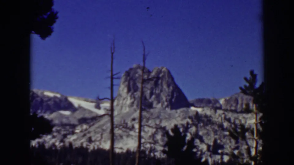 1939 Lake Tahoe home-movie frame showing a snow-dusted rocky ridge beneath blue sky, with pine silhouettes and bare trunks in the foreground.