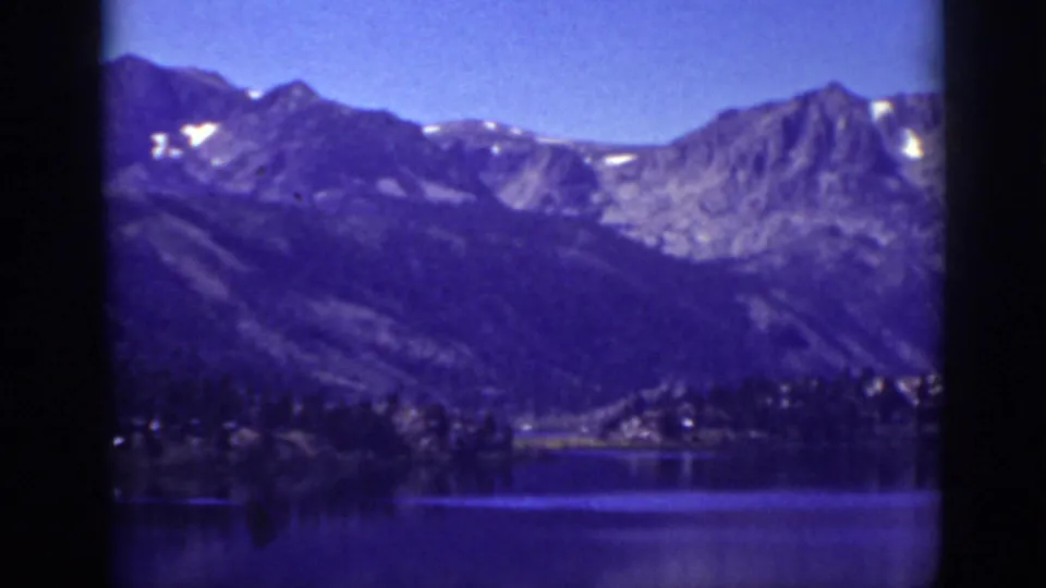 Late-1930s Lake Tahoe home-movie frame showing a quiet lake, wooded shore, and rugged mountains in saturated blue-purple color.