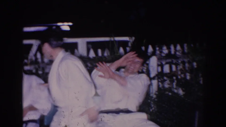 In 1966 El Monte home-movie footage, two men in white martial arts uniforms appear to prepare or spar near a fence under night lighting.