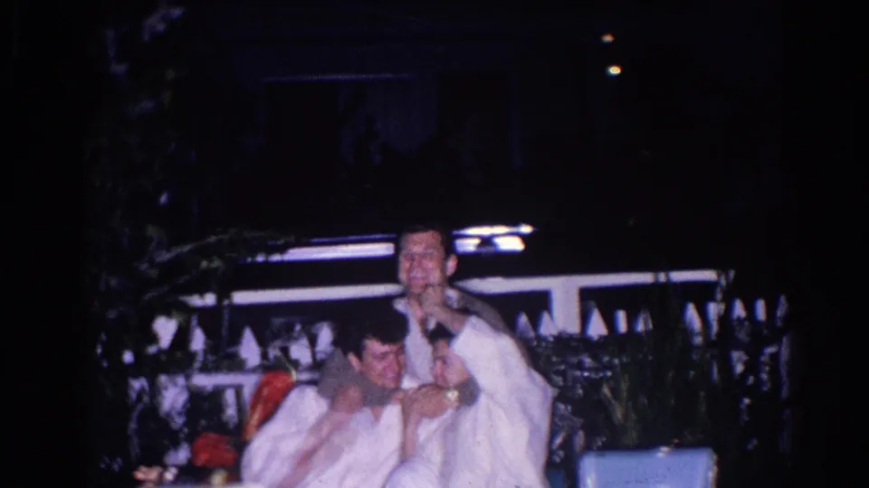 Two men in white uniforms sit together in the 1966 El Monte home movie, smiling near a fence in low night lighting.