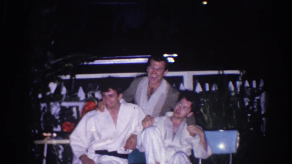 Three young men in martial arts uniforms laugh and pose together on a dimly lit El Monte patio in 1966.