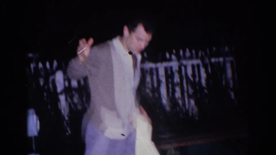An adult man in light clothing raises an arm during the El Monte nighttime backyard karate practice, with a fence behind him.