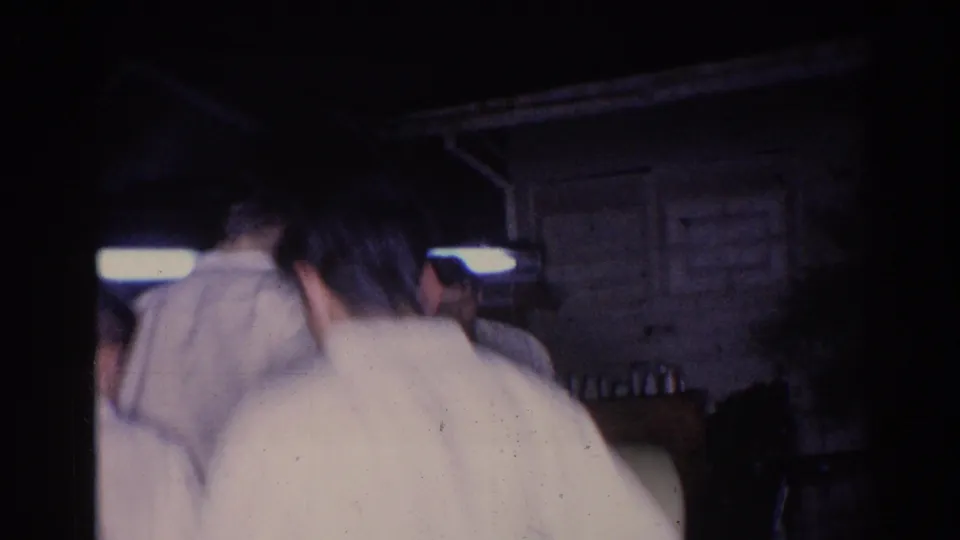 The camera trails participants in white gi from behind as they stand near a dimly lit exterior space in El Monte.