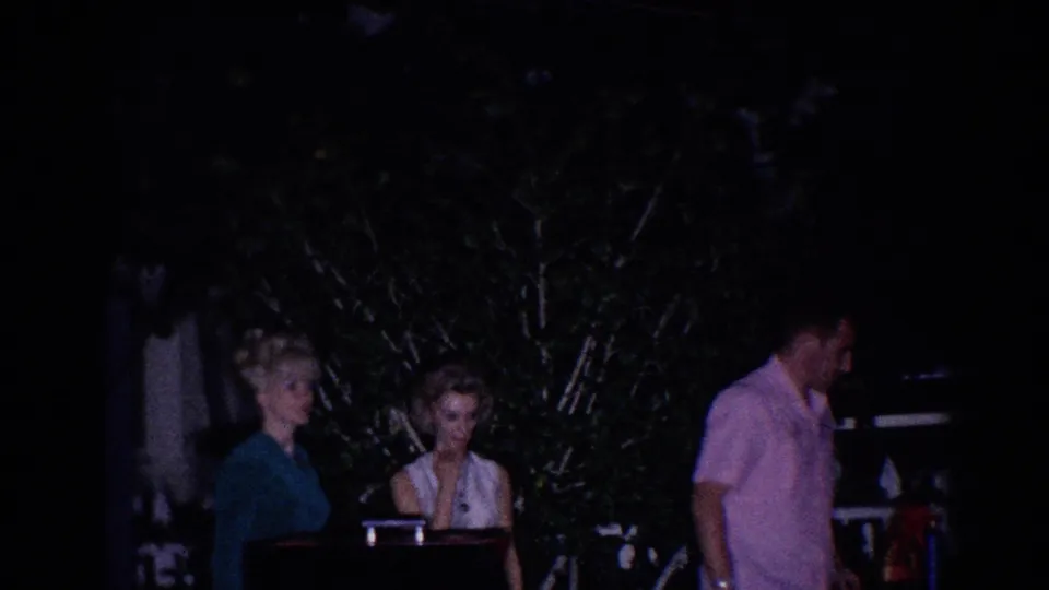 Adults gather outdoors at night in El Monte, with two women near a piano-like surface and a man in a pink shirt at the edge.