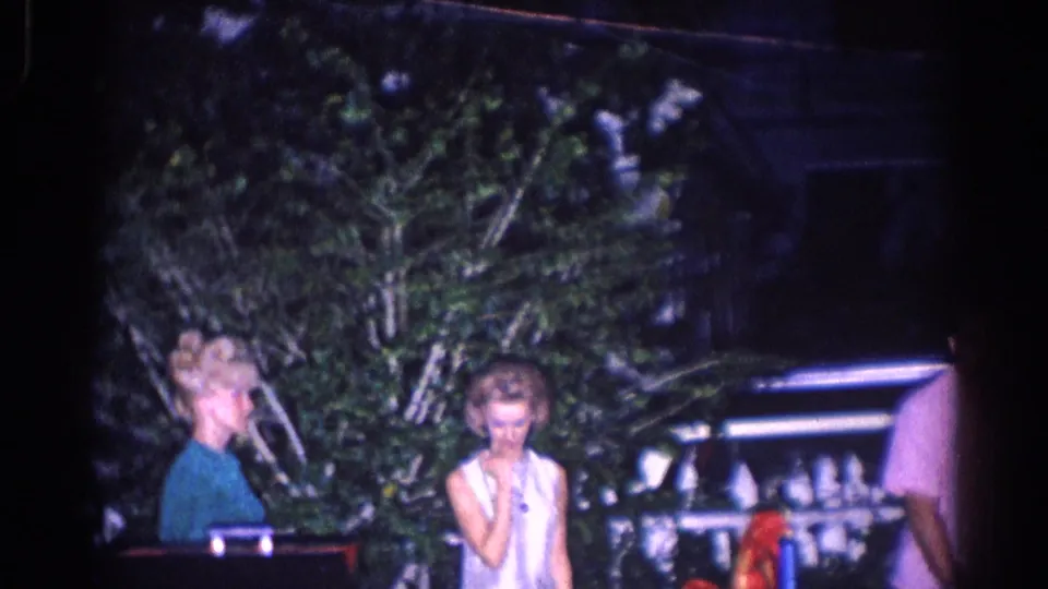 Two women at the 1966 El Monte gathering stand near trees and a piano-like object while another guest is partly visible.