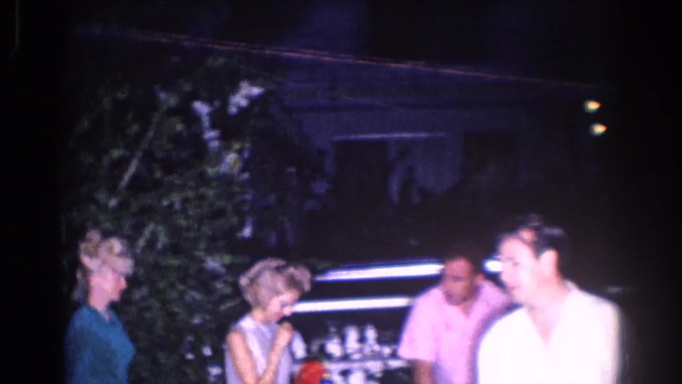Adults at a dim 1966 El Monte outdoor party mingle near trees, with a man in a white shirt crossing the foreground.