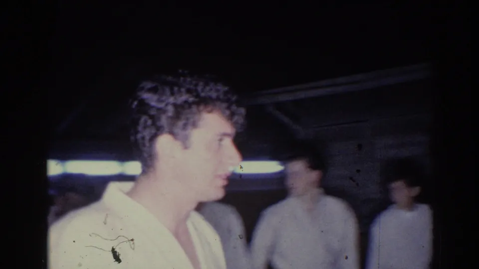 In 1966 El Monte home-movie footage, a man in a light robe-like garment stands in profile under dim artificial light.
