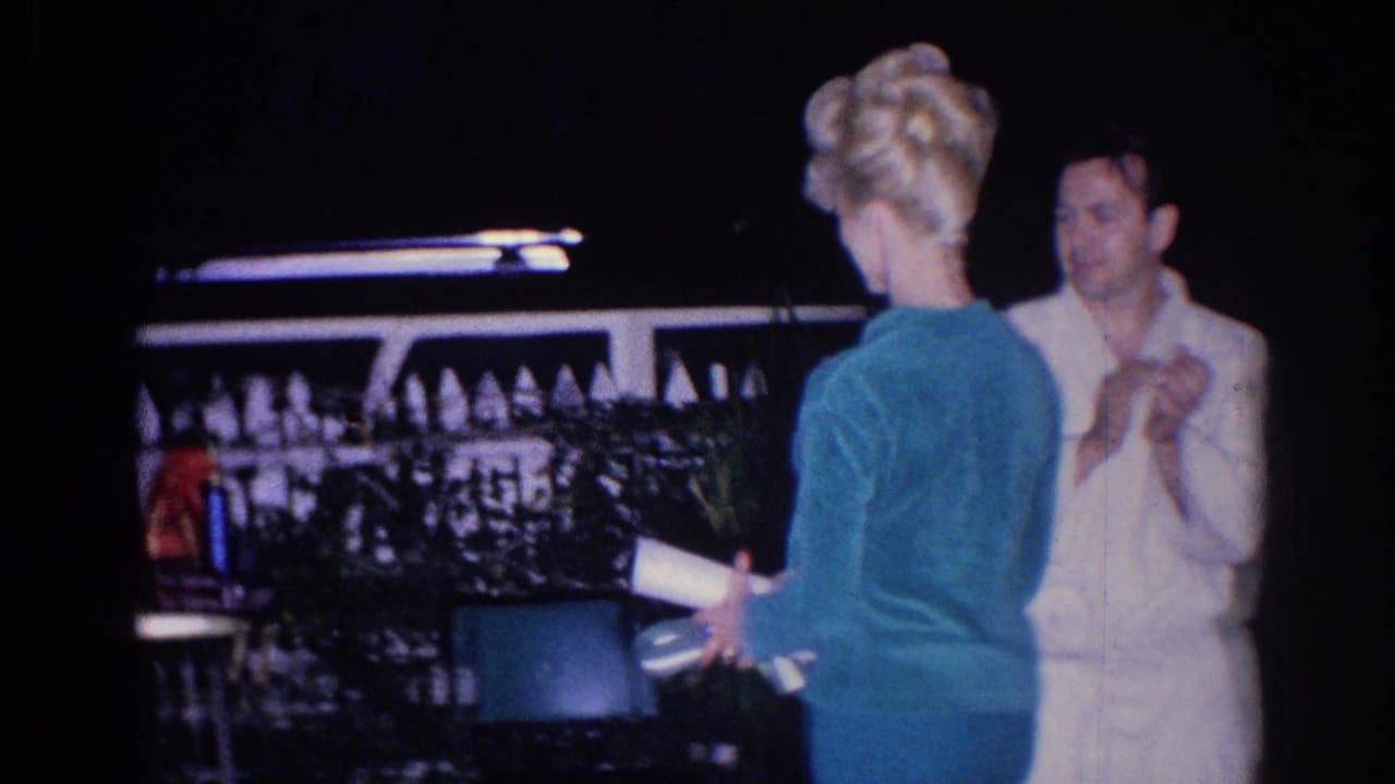 Adults in dress clothing talk outdoors at night near a white fence and greenery.