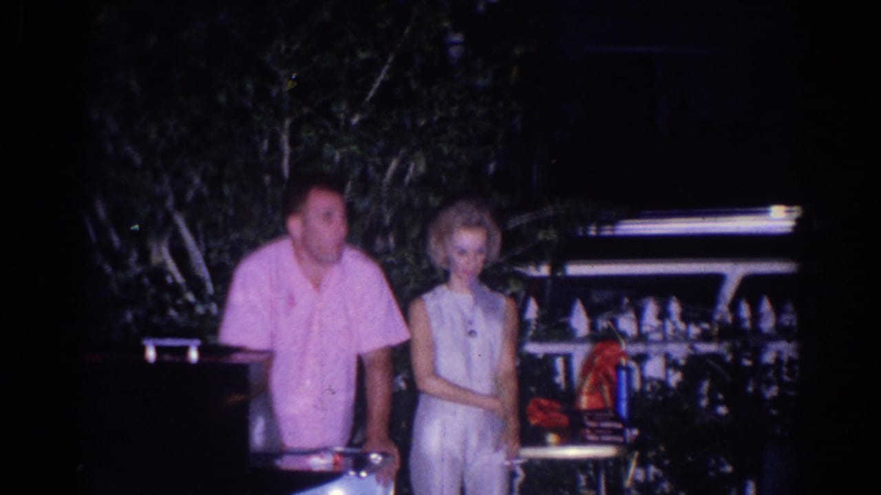 A man in a pink shirt stands near a woman in white beside outdoor furniture at night.