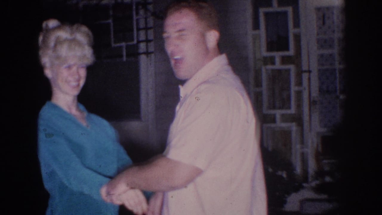 In 1966 El Monte home-movie footage, a man in a light shirt and a woman in a teal top hold hands near geometric glass doors.