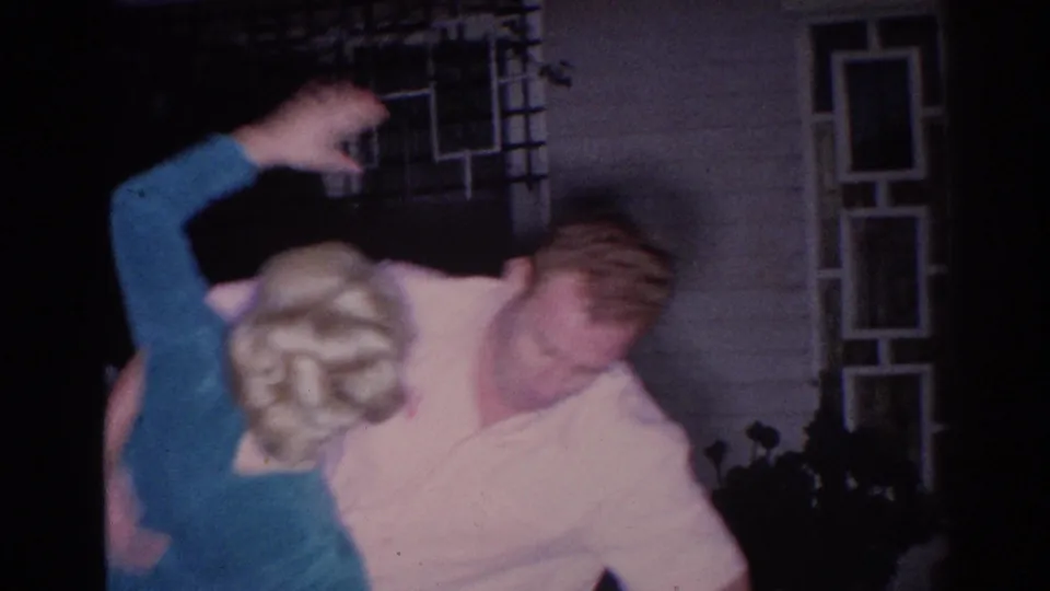 The couple moves into a playful embrace or lift outside a home, with the woman's arm raised and the doorway behind them.