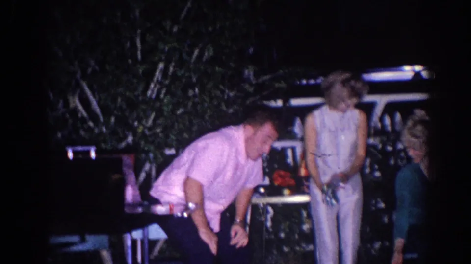 In the 1966 El Monte nighttime scene, the man in the pink shirt bends forward beside a woman in white near patio furniture.