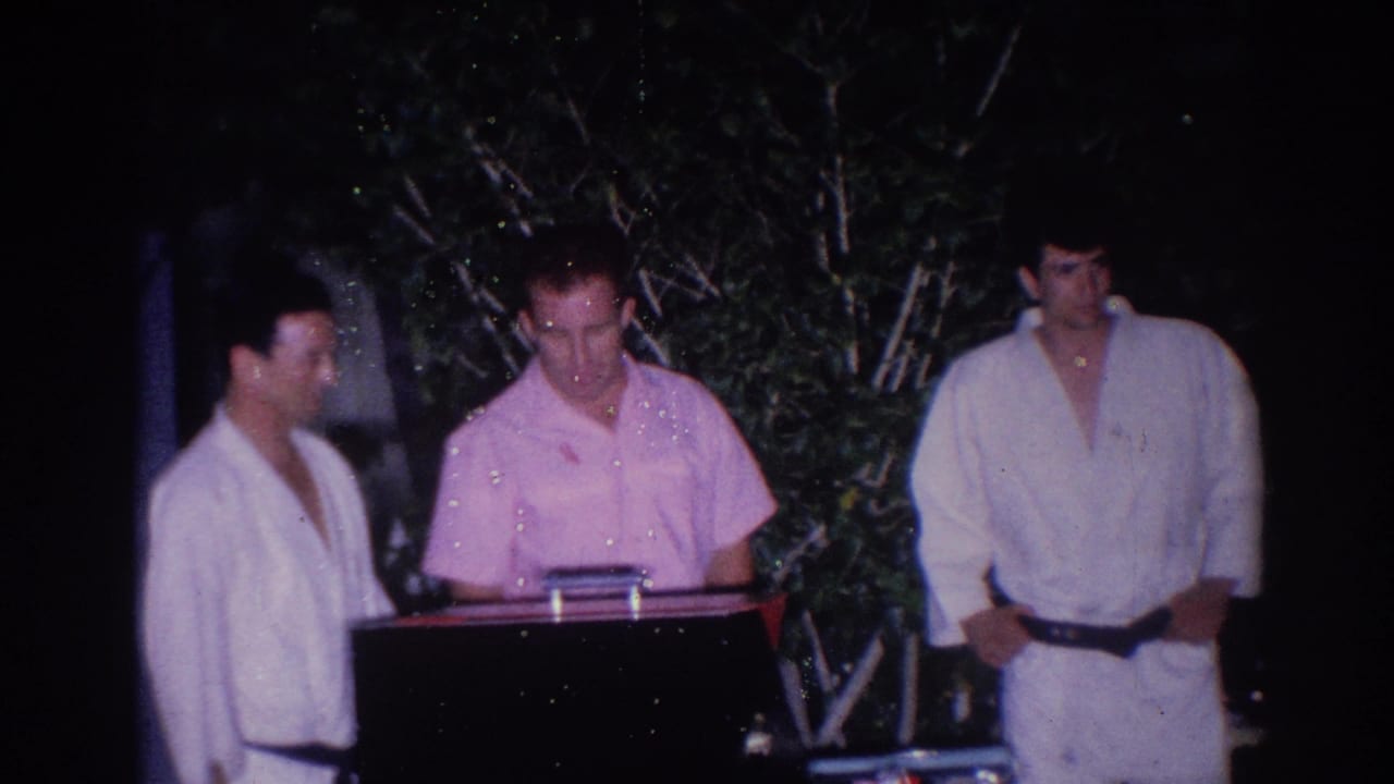 Two men in white uniforms stand near a man in a pink shirt at a small stand outdoors.