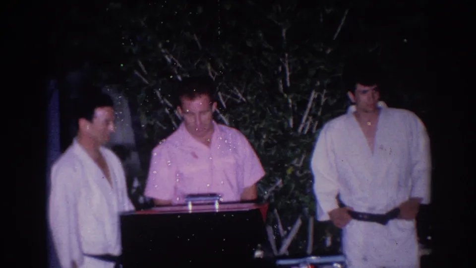 1966 El Monte frame with martial arts participants beside a judge or official at a stand during a nighttime test or demonstration.