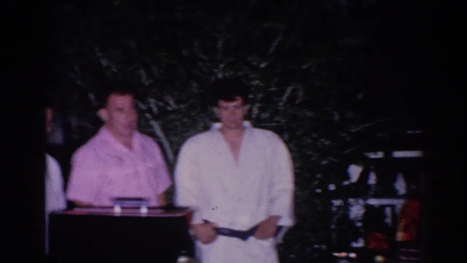 1966 El Monte home-movie frame showing a judge or official next to a uniformed martial arts participant under night lighting.