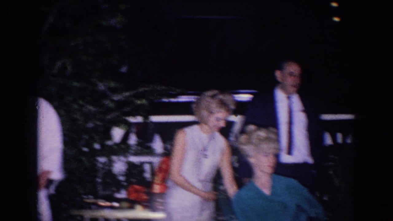 Adults at a dim outdoor gathering, including a woman in a light dress and a man in a dark suit near foliage and a vehicle.