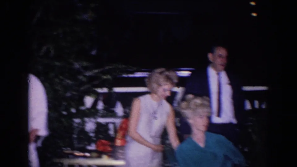 1966 El Monte home-movie frame showing adults at a nighttime outdoor gathering, with a woman in a light dress, a suited man, foliage, and a parked vehicle.