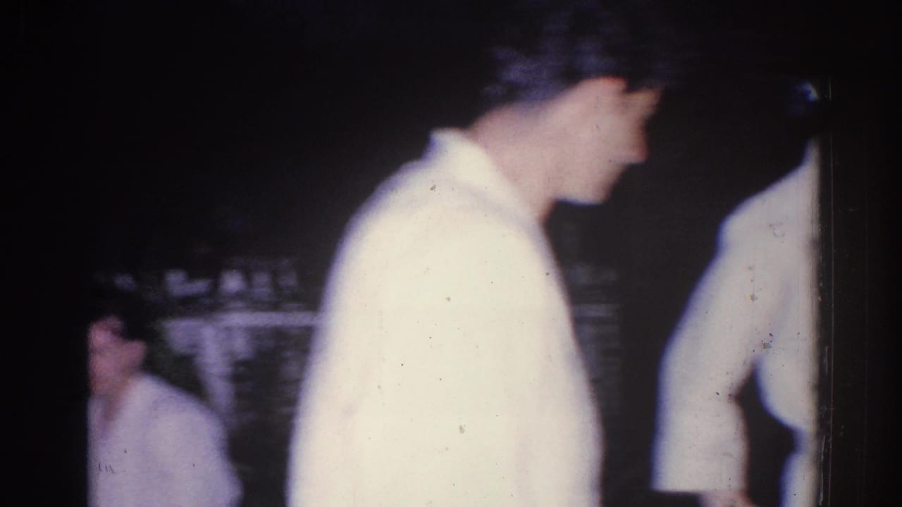 Young man in a white shirt stands close to the camera while other adults are partly visible in the dim background.