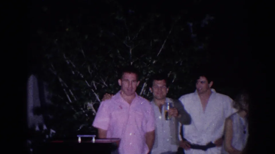 1966 El Monte home-movie frame showing three men at an outdoor nighttime gathering, including one holding a drink, with foliage behind them.