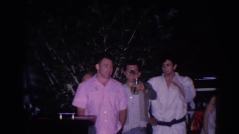 In 1966 El Monte home-movie footage, three adult men stand together at a nighttime backyard gathering, one holding a drink.