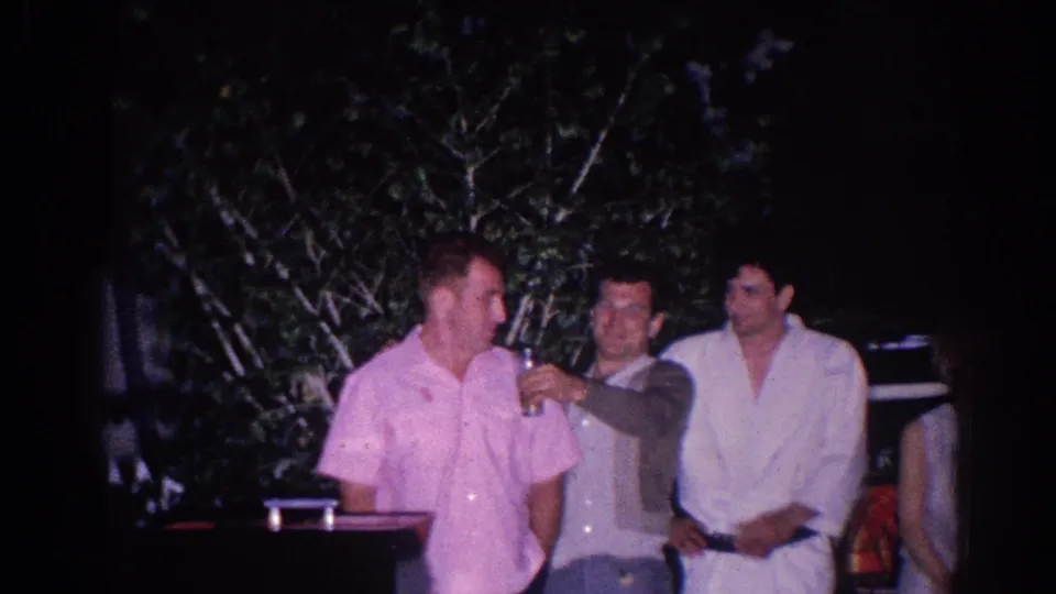 Three adult men socialize outdoors at night in El Monte, with a drink raised between them and foliage behind them.