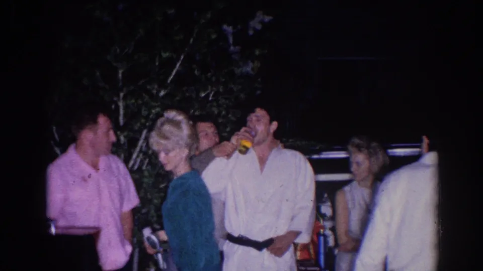 In 1966 El Monte home-movie footage, adults and judo participants stand under night lighting while one man in a white gi drinks from a yellow cup.