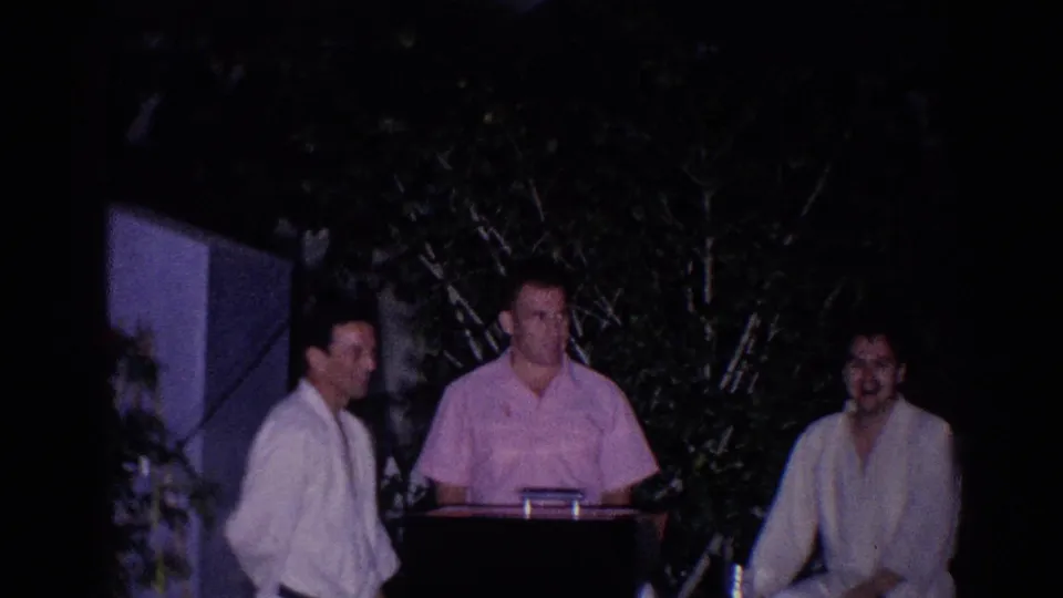 Three adults stand in a dark outdoor gathering in 1966 El Monte, with trees behind them and low-light 8mm grain.