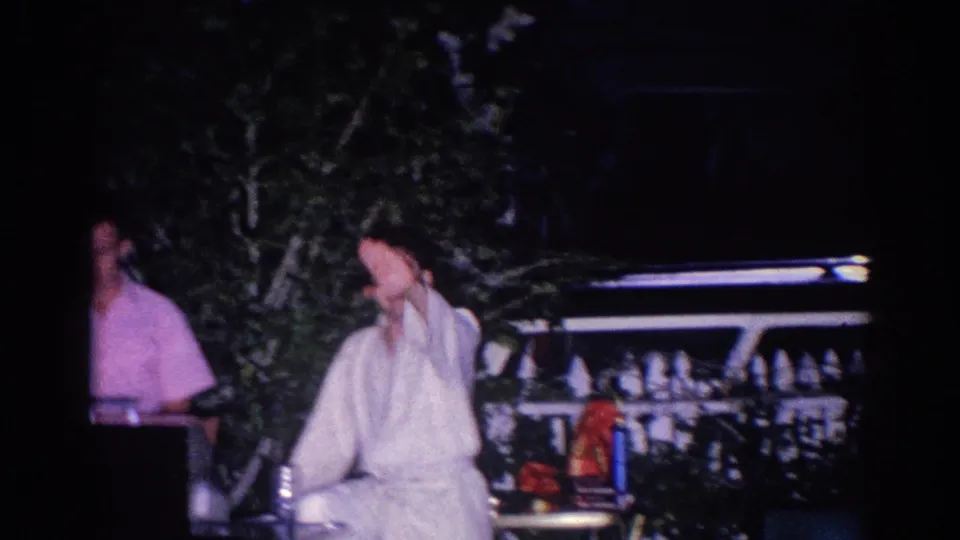 A seated person in white clothing gestures toward the camera during a nighttime 1966 El Monte outdoor gathering.
