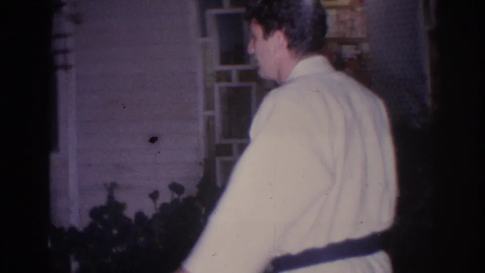 A man in a white gi stands with his back to the camera near the exterior wall and windows of a house in the 1966 El Monte footage.