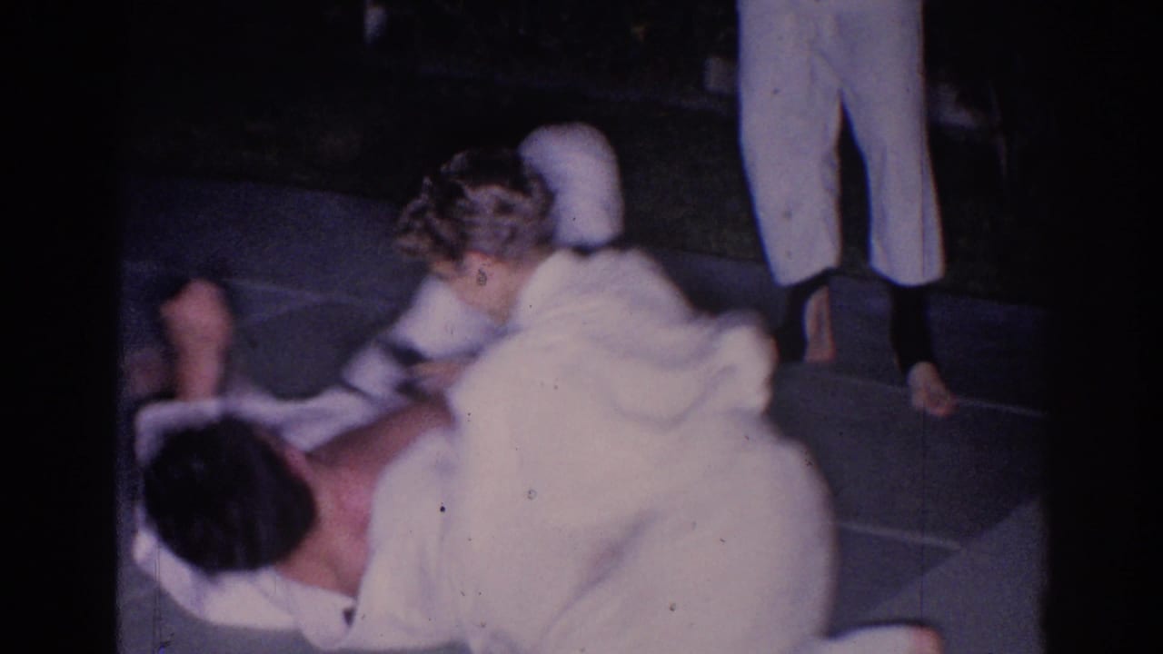 Two people in white karate gi uniforms grapple on a paved surface at night.