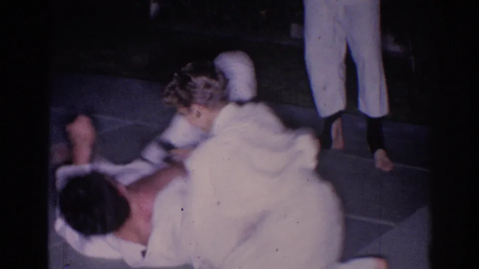 1966 El Monte home-movie frame showing two gi-clad martial arts participants in a low grappling position on pavement at night.
