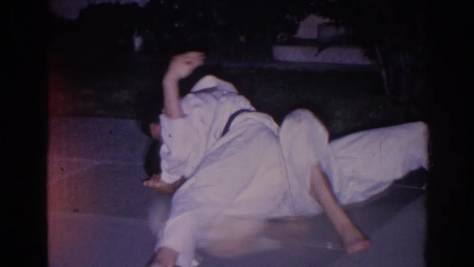 1966 El Monte home-movie frame with two gi-clad martial arts participants on the pavement in a grappling or rolling practice pose.