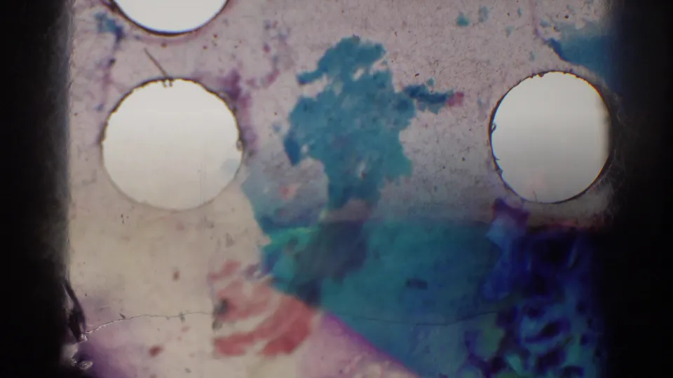 8mm film artifact frame showing sprocket holes, stained emulsion, and abstract blue and red marks rather than a clear scene.