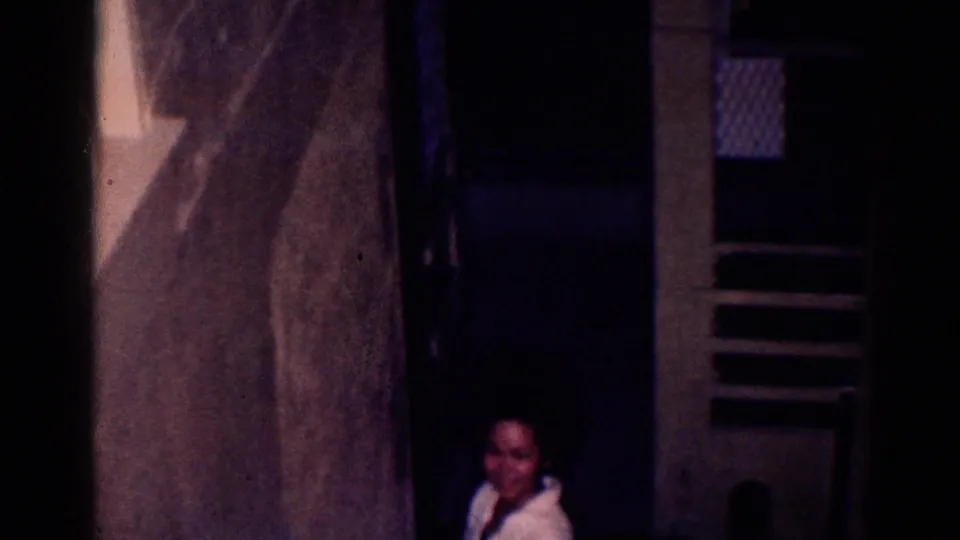 1966 El Monte home-movie frame showing a dim interior, a woman near a doorway, and shadowed architectural details.