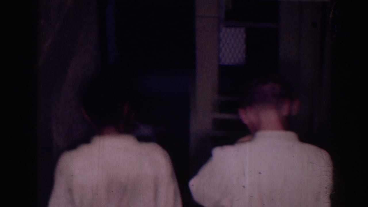 Two athletes in white uniforms walk toward a doorway in a dim room.