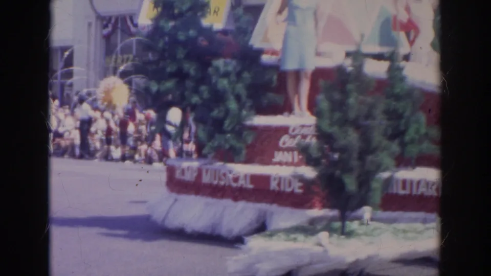 1966 Norfolk parade view of a passing decorated float with trees, standing people, crowd spectators, and partial musical ride lettering.
