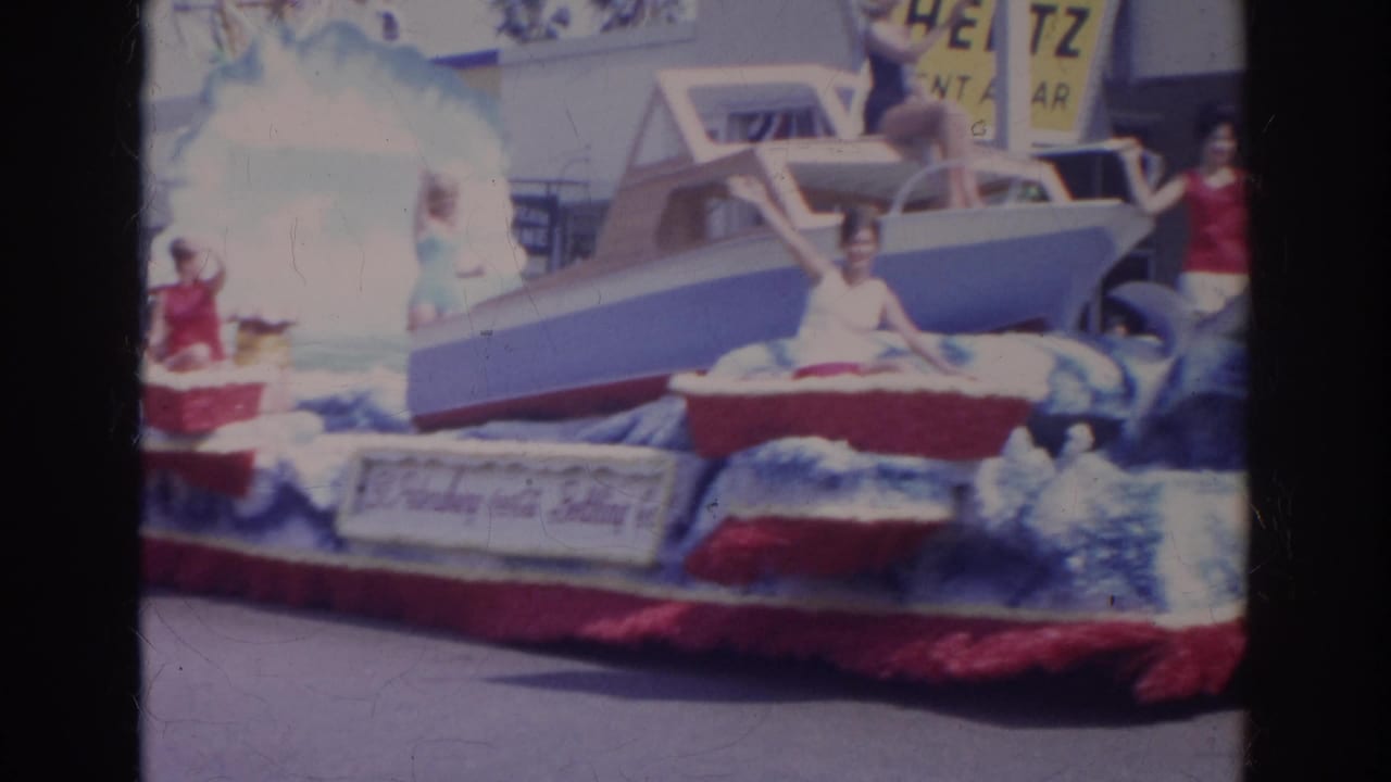 Red, white, and blue parade float with model boat and women waving on a city street.