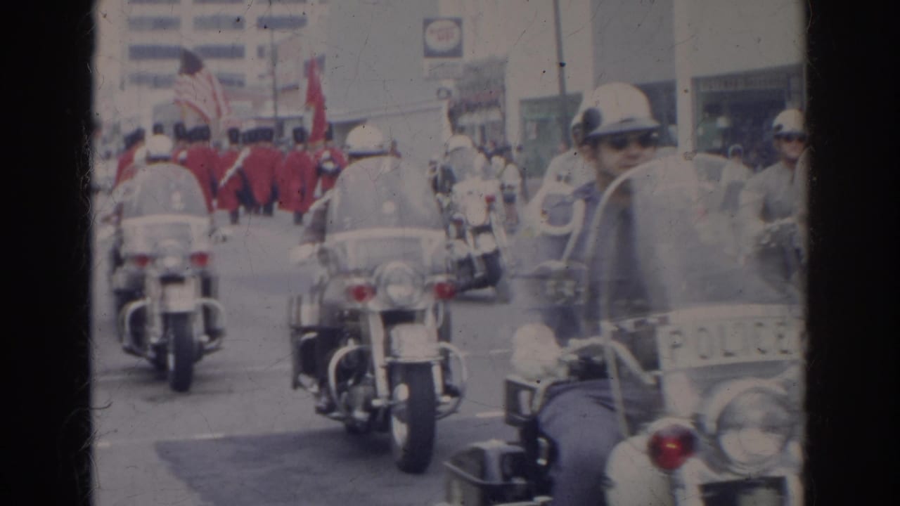 Police motorcycles lead a parade with red-uniformed marchers behind them on a city street.