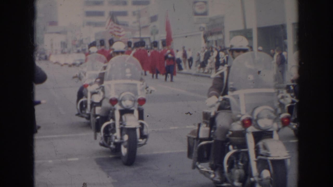 Motorcycle police ride toward the camera with a marching group and flags behind them.