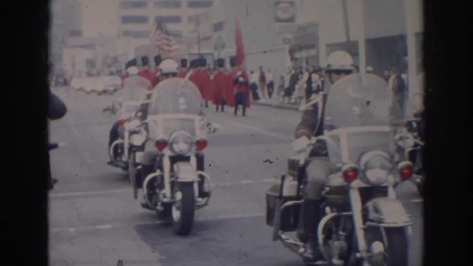 Motorcycle officers escort a 1966 Norfolk parade as red-coated marchers and flags approach along a downtown street.