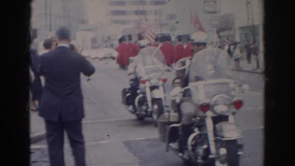 A spectator photographs the 1966 Norfolk parade while motorcycle police and red-uniformed marchers continue down the street.