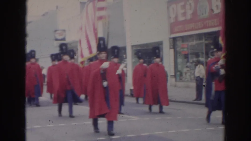 Red-coated marching band members with tall black hats proceed through a Norfolk city street during the 1966 parade.