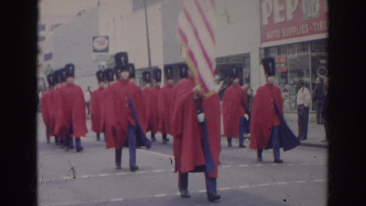 Red-caped marching band members move down a city street with an American flag.