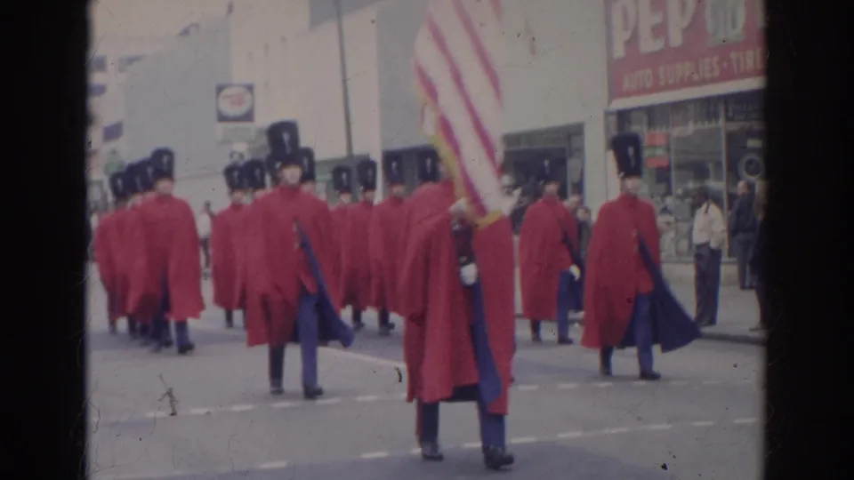 In 1966 Norfolk, Virginia, red-caped parade marchers in tall black hats pass storefronts while one carries an American flag.