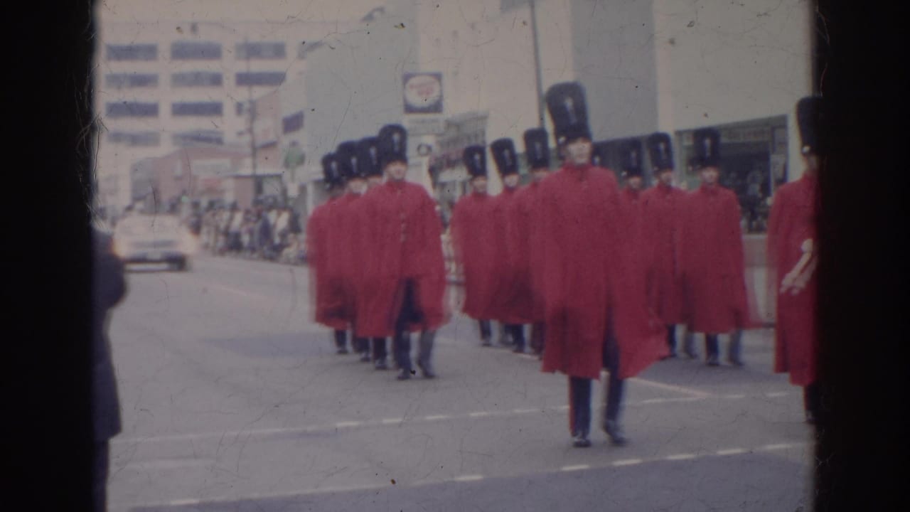Red-caped marchers in tall black hats advance through the Norfolk Fourth of July parade with spectators and cars in the background.