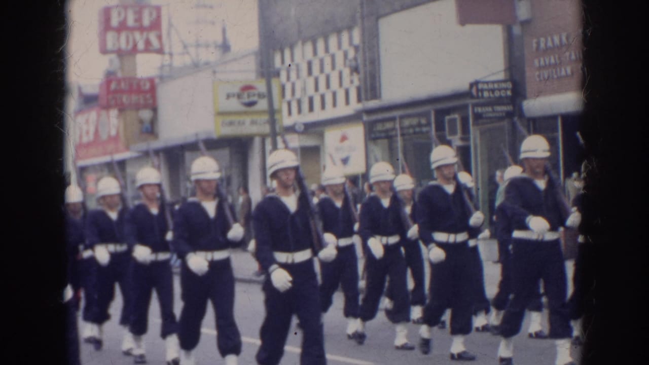 Sailors in dark uniforms and white helmets march past storefront signs on a city street.