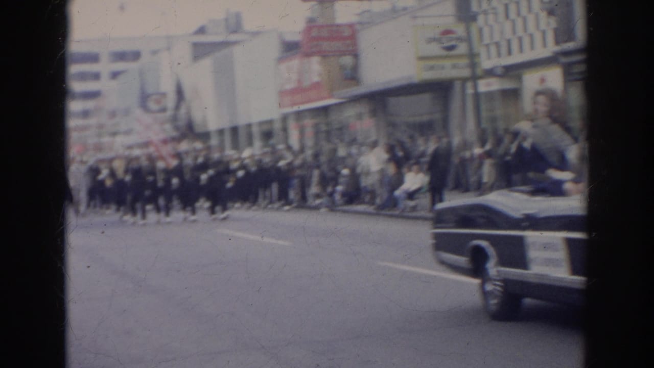 Street parade with a convertible, marching band, spectators, and storefronts.